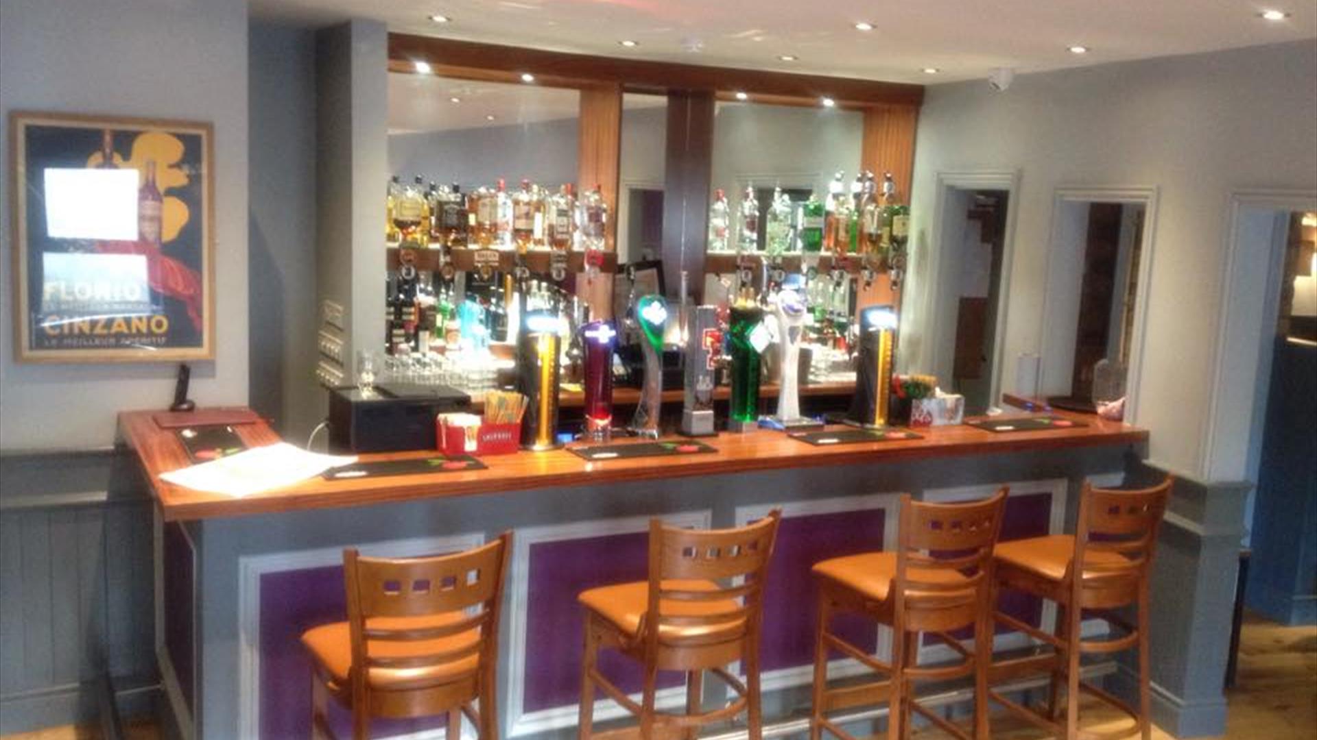 Image shows bar area with 4 bar stools on wooden bar bench and bar is painted a soft lilac colour with recessed lighting overhead.