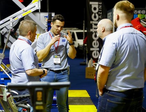 Image shows 4 men standing chatting at a trade show, with machinery & trade equipment in the background.