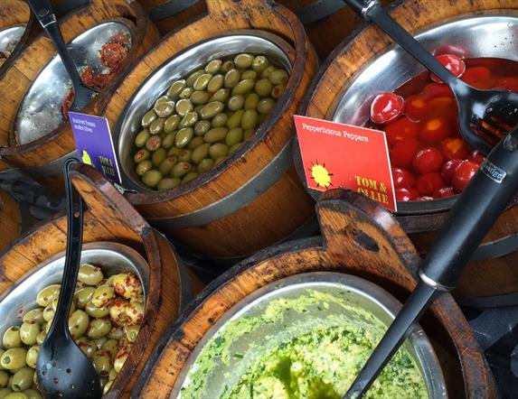 Image shows wooden barrels of different foods such as olives at the market