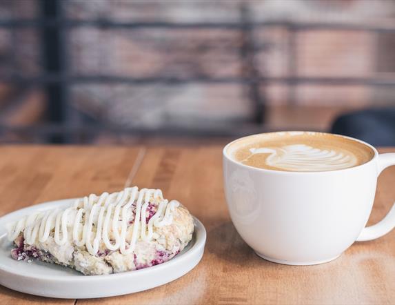 Image shows a white cup of latte and white side plate with a  traybake covered in icing.