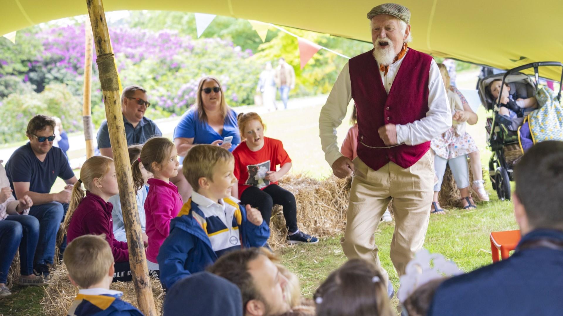 Image shows man in a purple waist coat with cap dancing with a group of children looking on