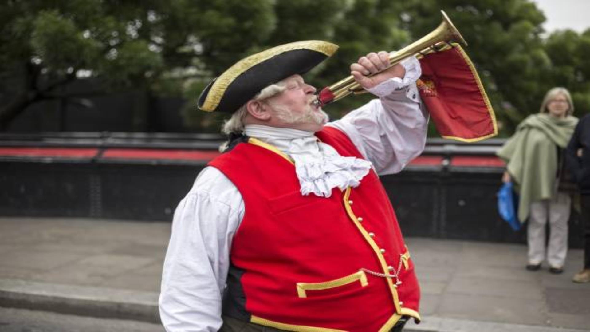 Image shows Town Crier dressed in traditional dress with red waistcoat and black hat.