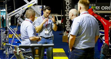 Image shows 4 men standing chatting at a trade show, with machinery & trade equipment in the background.