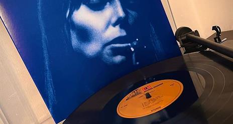 Picture of Joni Mitchell on the cover of her album in the background with a vinyl record on record player to the fore