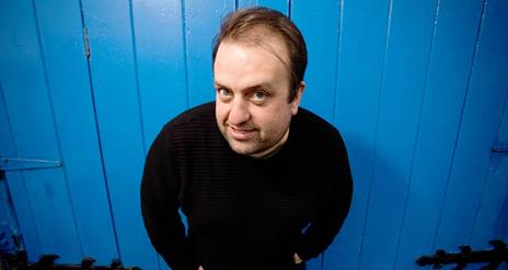 Image is of the comedian Karl Spain smiling and standing in front of a blue wooden door