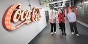 Image is of a lit up Coca-Cola sign and visitors looking at it