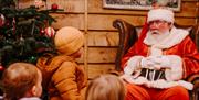 Image shows Santa sitting in his armchair wearing white gloves with his hands clasped, chatting to three young children