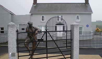 Image is of Harry Ferguson bronze statue leaning on gate  in front of his family home