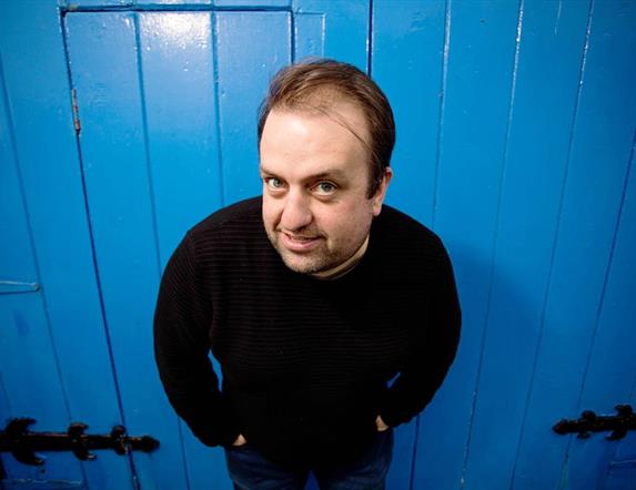 Image is of the comedian Karl Spain smiling and standing in front of a blue wooden door