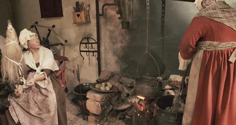 Image is a scene from a weaver's cottage where women are cooking on a fire and one woman is weaving linen